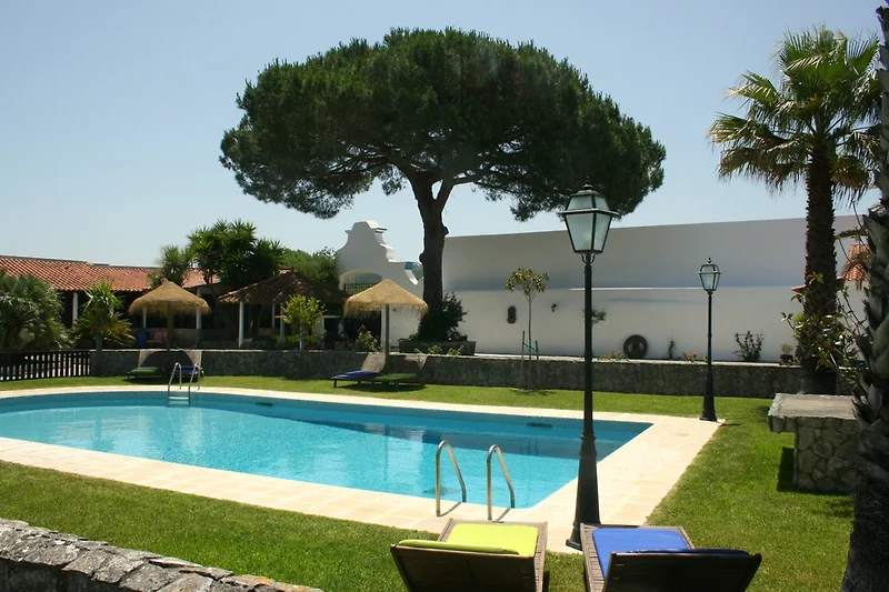 Zwembad met mensen die ontspannen en genieten van het water. Sommigen zwemmen, terwijl anderen op ligstoelen zonnen. Er zijn parasols die schaduw bieden en het gebied rond het zwembad is betegeld en schoon. Het water is helder en nodigt uit tot een verfri