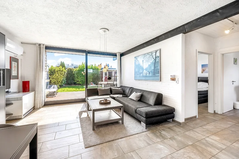 Wohnzimmer mit Couch, Fernseher und Tageslicht.