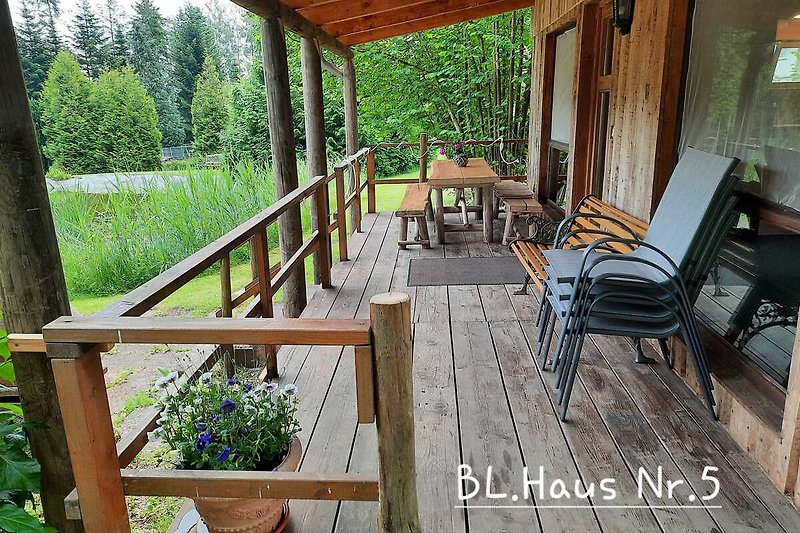Holzhaus mit Terrasse, Tisch und Stühlen, umgeben von Pflanzen und Natur.