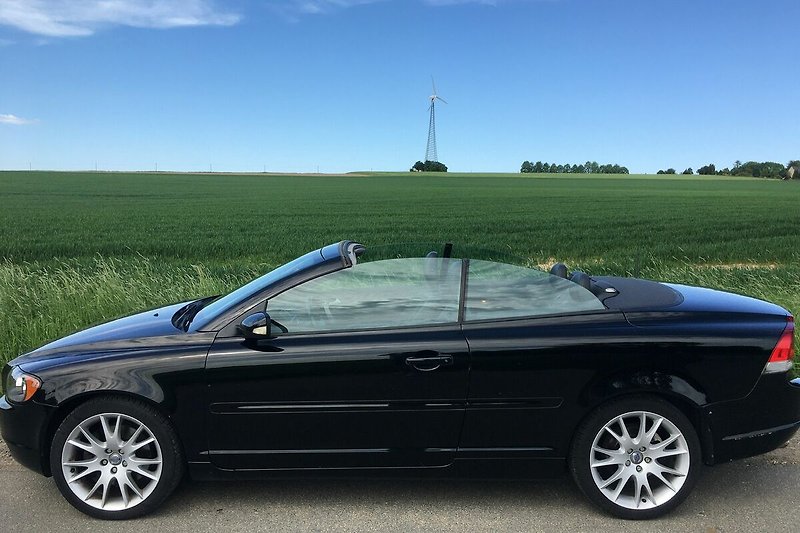 Inklusive Leihwagen Volvo C 70 Cabriolet Vollausstattung ageno vor blauem Himmel und grüner Landschaft.