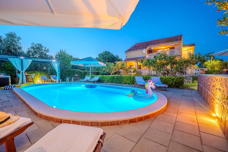 Beleuchteter Poolbereich mit Palmen und Gartenmöbeln bei Nacht.