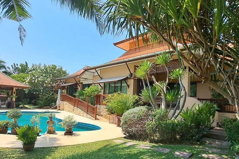 Schwimmbad mit Sonnenschirmen und Liegestühlen, umgeben von Palmen und Grünflächen.