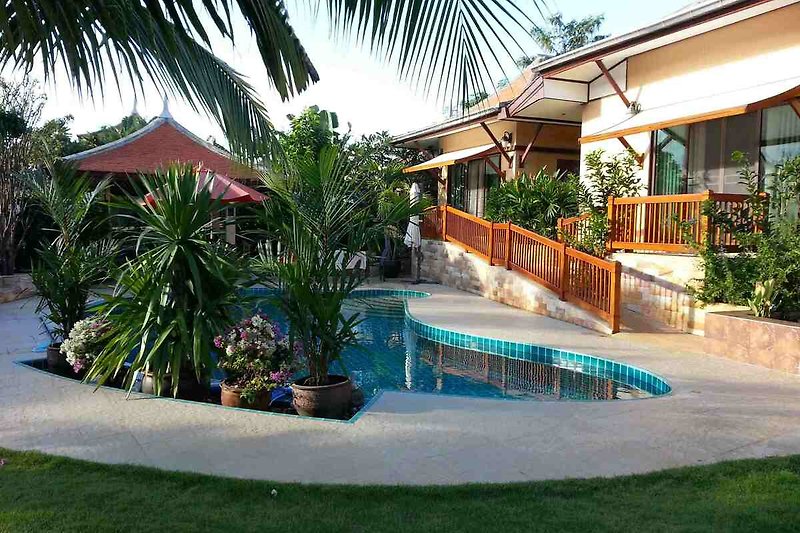 Schwimmbad mit Sonnenschirm und Liegestuhl unter Palmen.