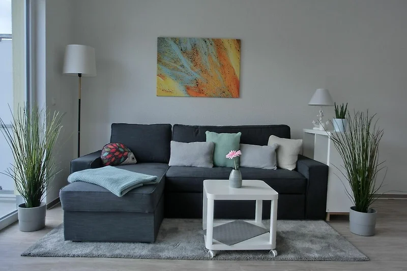 Soggiorno con divano grigio e cuscini colorati, tavolino in legno, tappeto beige e pareti bianche con decorazioni moderne.