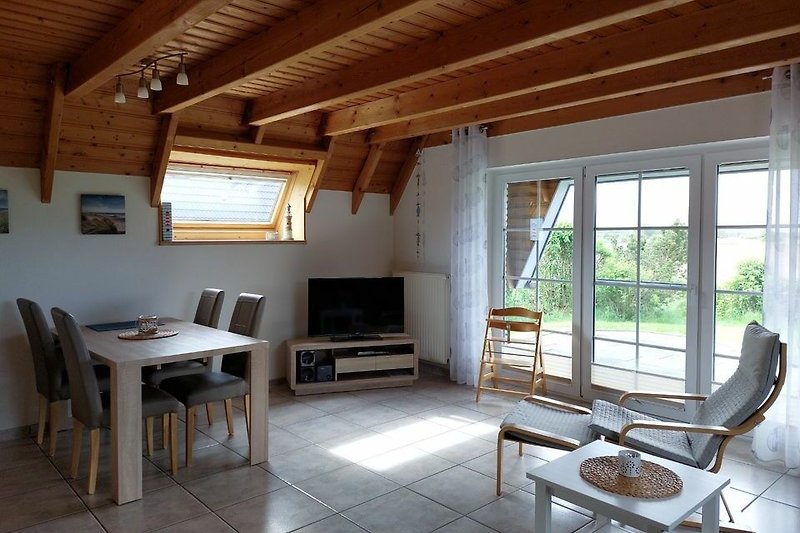 Gemütliches Wohnzimmer mit Holzmöbeln, Couch und Pflanzen. Große Fenster sorgen für viel Tageslicht.