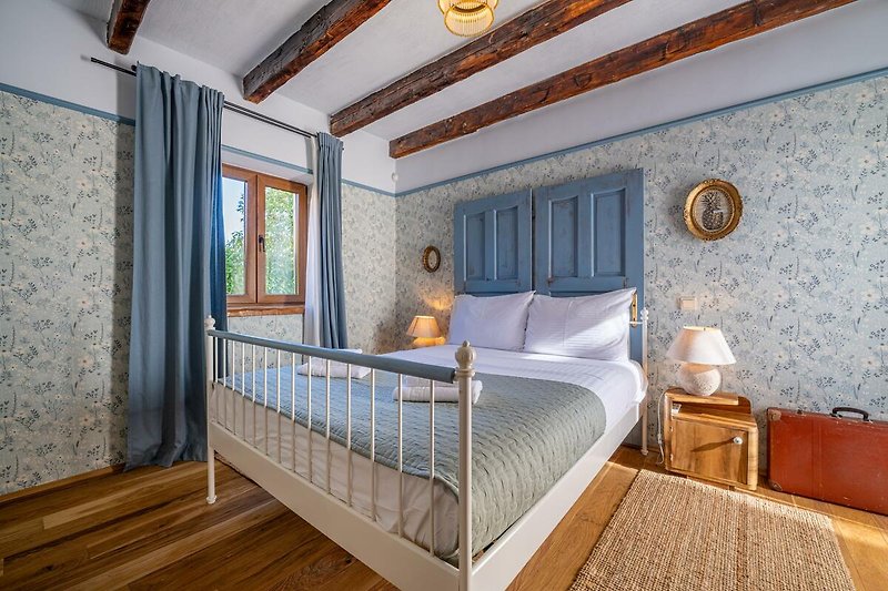 Gemütliches Schlafzimmer mit Holzbett, Fenster mit Vorhängen und gemütlicher Bettwäsche.