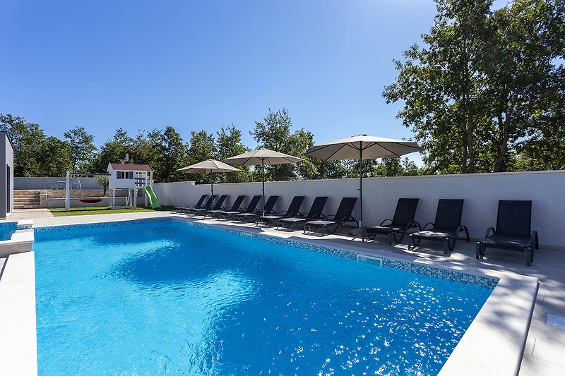 Predivan pogled na bazen s plavom vodom, okružen zelenilom i udobnim vanjskim namještajem.