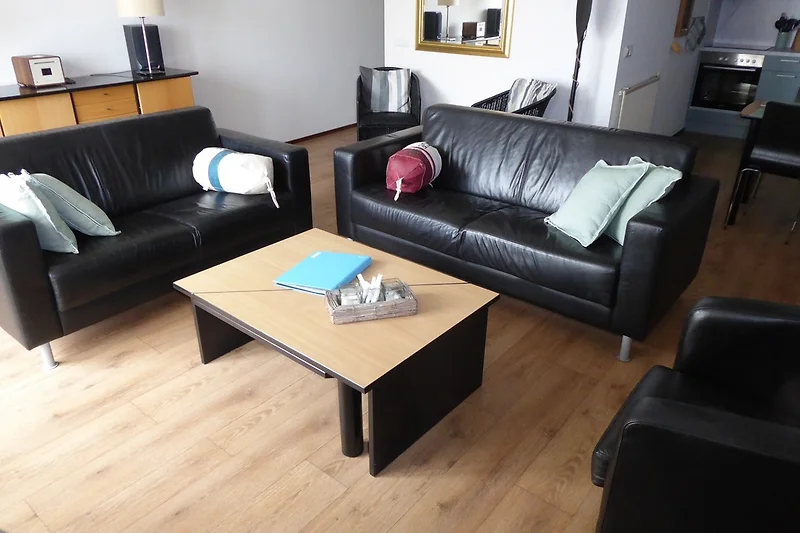 Zithoek met een moderne grijze bank en kleurrijke kussens in een lichte woonkamer met grote ramen en een groen uitzicht.