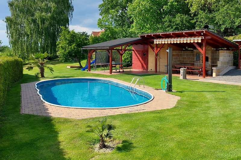 Schwimmbad, Garten, Haus und Außenmöbel unter blauem Himmel.