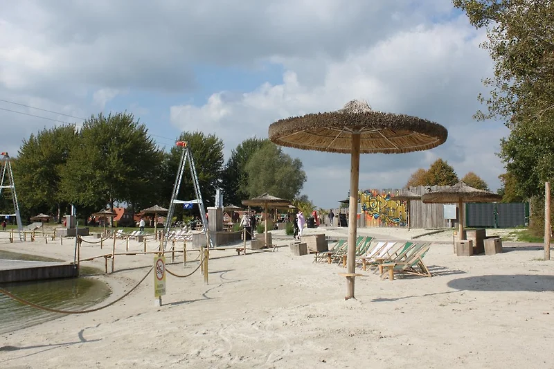 Strandbar aan het meer met zandstrand, parasols, ligstoelen en een houten loopbrug. Bezoekers genieten van drankjes en ontspannen in de zon.