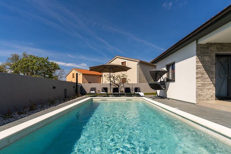 Luxuriöses Anwesen mit Pool, Sonnenliegen, grünen Pflanzen und blauem Himmel.