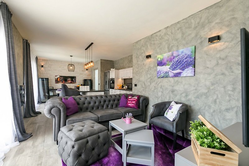 Gemütliches Wohnzimmer mit lila Couch und violetten Akzenten. Tisch, Stühle und Bilderrahmen vervollständigen das Interieur.