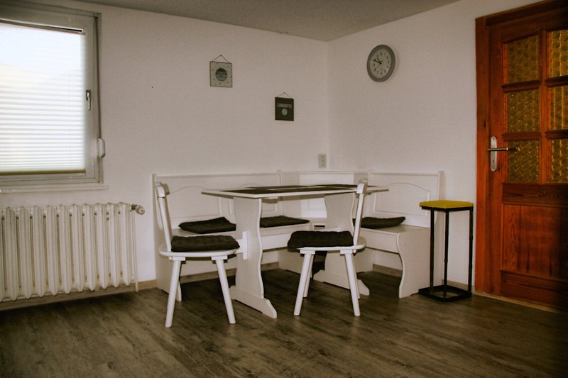 Küchentisch mit Stühlen, Uhr an der Wand, Holzfenster.