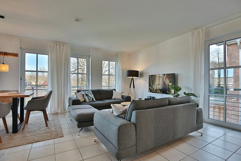 Woonkamer met een grote grijze bank, een witte koffietafel en een groot raam met gordijnen. Er is een televisie aan de muur en verschillende planten in de hoek van de kamer.