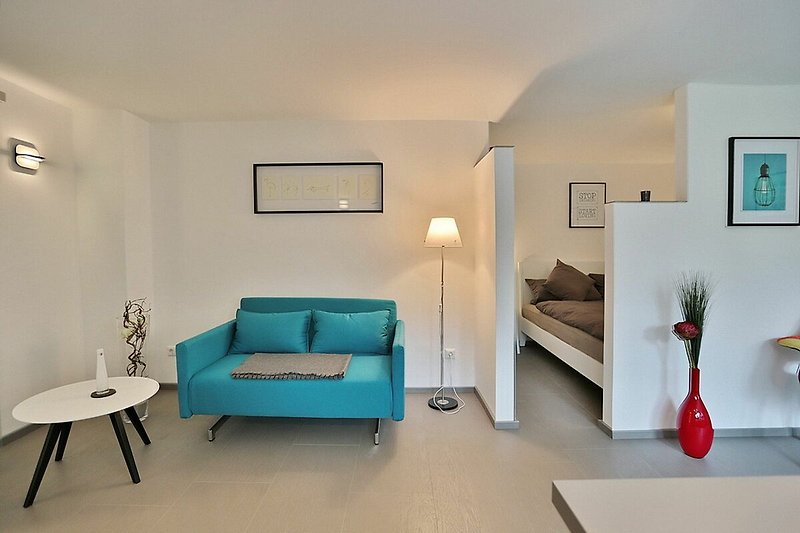 Woonkamer met een grote grijze hoekbank, een laag wit koffietafeltje en een groot flatscreen-tv aan de muur. Er is een groot raam dat veel natuurlijk licht binnenlaat. De vloer is van licht hout en er ligt een groot, zacht vloerkleed.