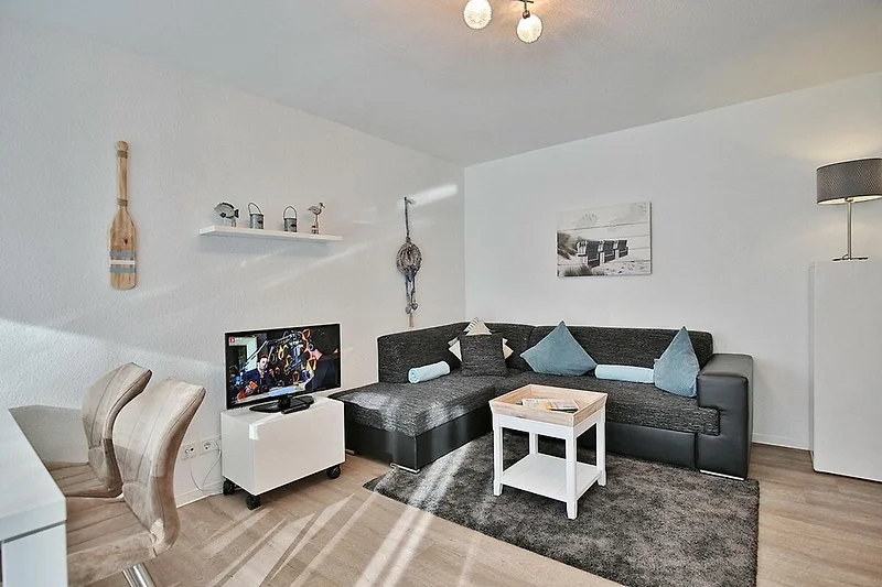 Salon avec un canapé gris, une table basse en bois et une grande fenêtre avec des rideaux blancs. Des coussins colorés sont dispersés sur le canapé, et des plantes vertes sont placées à différents endroits de la pièce. Un tapis à motifs se trouve au centr