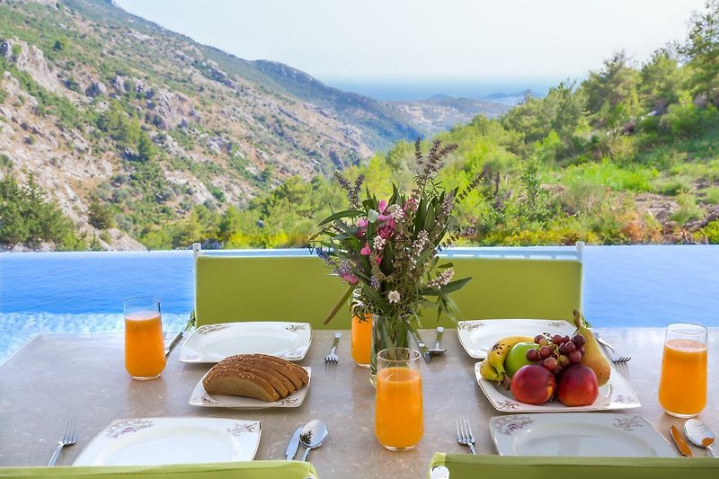 Blick auf Berge, Frühstückstisch im Freien, Blumen und Obst. Gemütliche Atmosphäre.