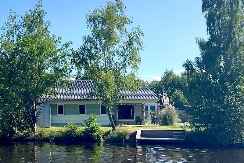 Haus am See mit Spiegelung im Wasser und grüner Umgebung.