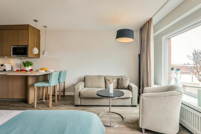 Soggiorno con divano grigio e cuscini colorati, tavolino in legno e tappeto beige. Una grande finestra lascia entrare molta luce naturale.
