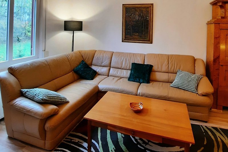 Gemütliches Wohnzimmer mit brauner Couch, Tisch und Kunst an der Wand. Helle Beleuchtung durch das Fenster.