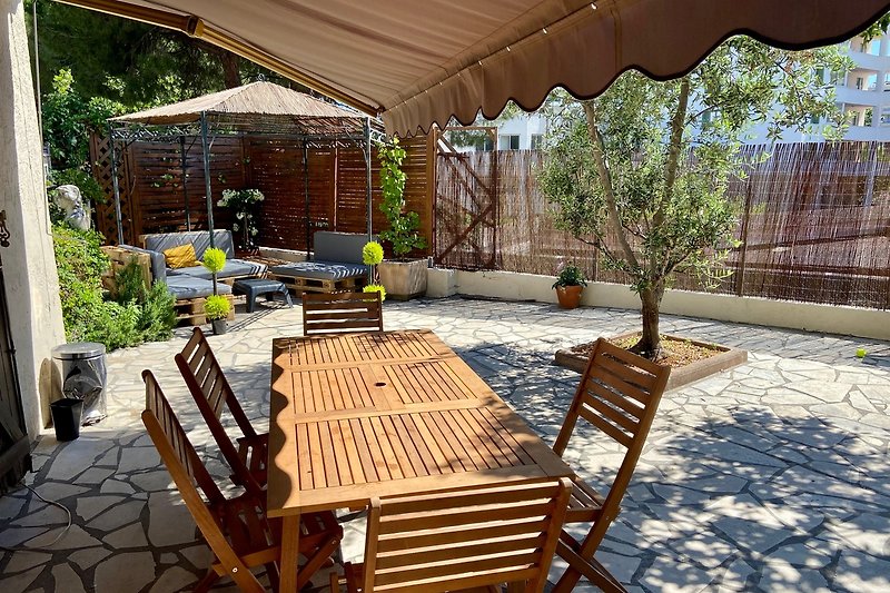 Une terrasse en bois avec des meubles d'extérieur, une table et des chaises, entourée de plantes et offrant de l'ombre.
