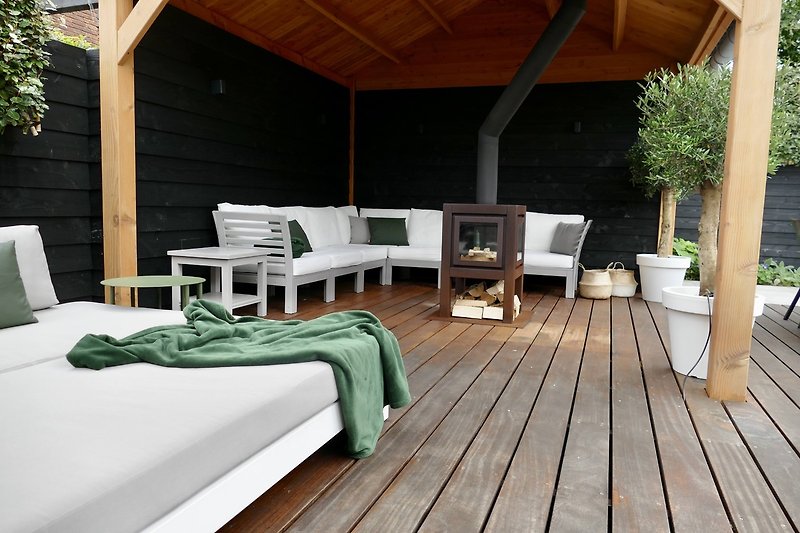 Zonnedek met pararol en loungeset naast de overkapte veranda met houtkachel