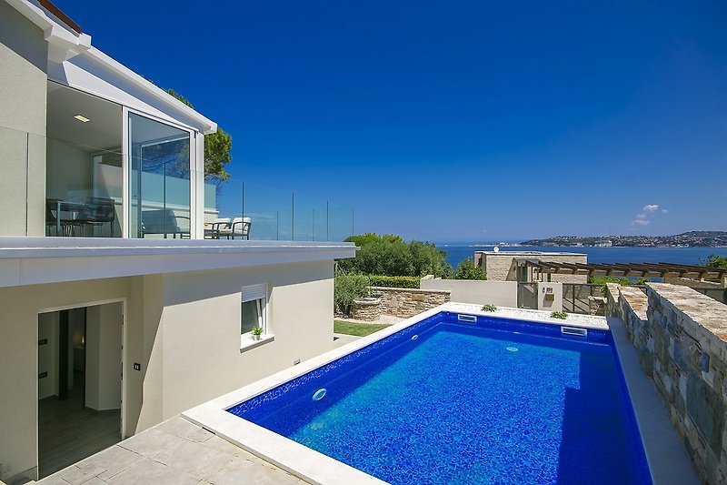 A stunning seaside villa with a pool, surrounded by lush greenery and outdoor furniture.