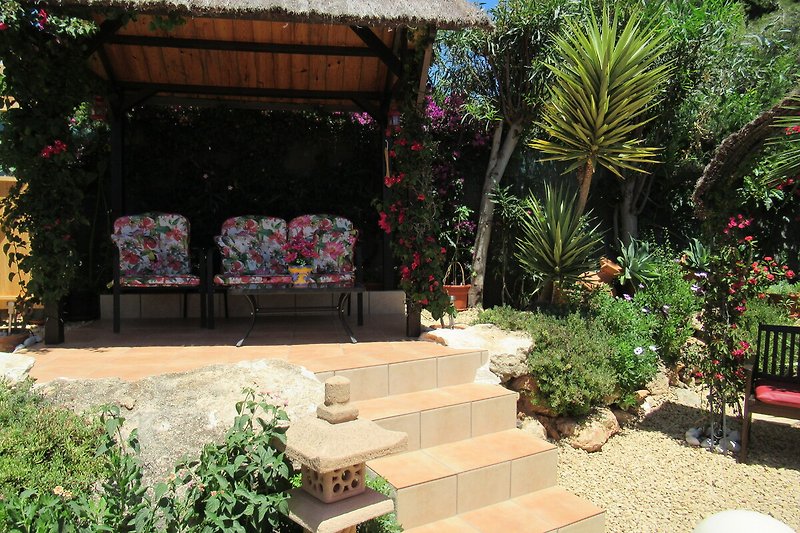 A picturesque garden with colorful flowers, outdoor furniture, and a charming pergola. Behind the pool area.