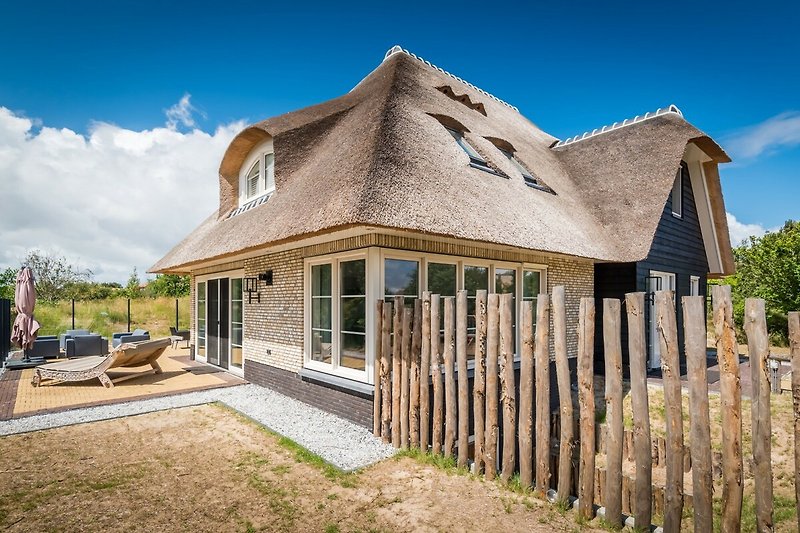 Ein charmantes Landhaus mit einem strohgedeckten Dach und einem malerischen Garten, umgeben von einer ländlichen Landschaft und einem Zaun.