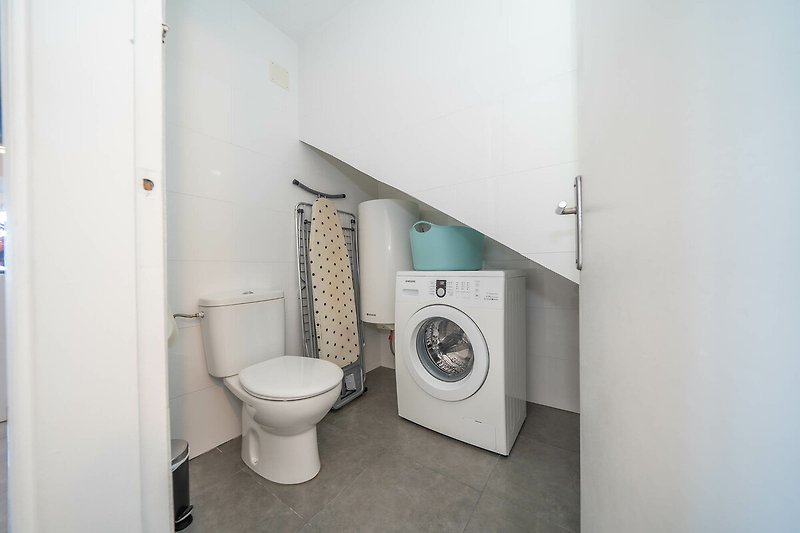 A nappali mellett található egy WC, ahol a mosógép, a szárítóállvány és a vasalóasztal is található.