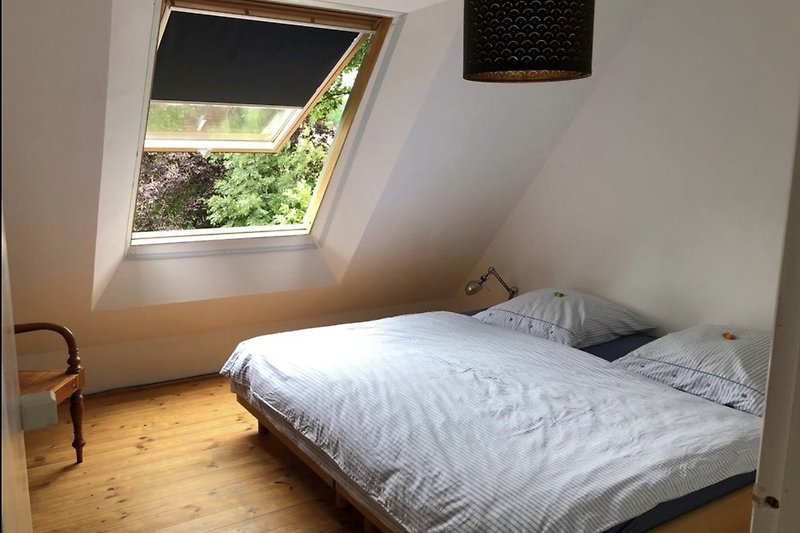 Schlafzimmer mit hellem Bett, Holzmöbeln und großem Fenster.
