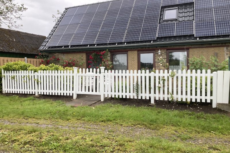 Haus mit Solarpanelen, Holzzaun, Garten und blühenden Sträuchern.