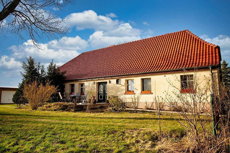 Ländliches Bauernhaus mit Garten, Bäumen und Wolken am blauen Himmel.