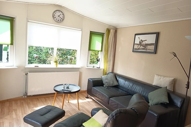 Wohnbereich mit Sofa, Sessel, Couchtisch, TV und großen Fenstern.