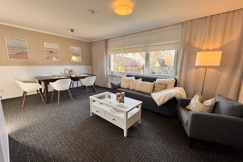 Wohnbereich mit Sofa, Sessel, Couchtisch, Pflanzen und großem Fenster.