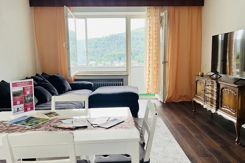 Wohnbereich mit Sofa, Tagesbett, TV, Esstisch und großen Fenstern.