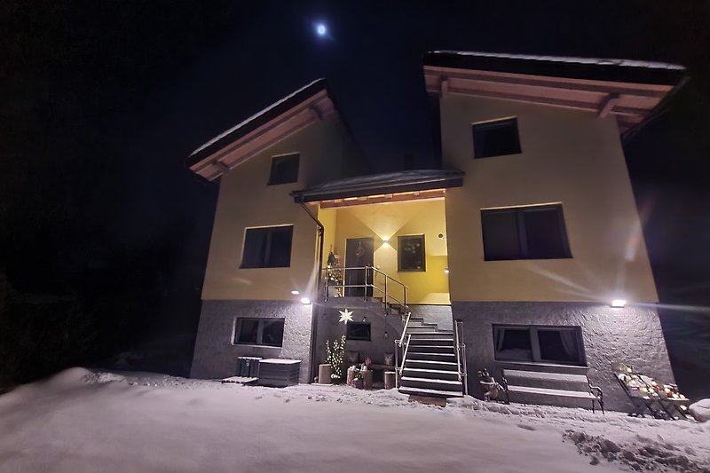 Gemütliches Haus mit winterlicher Landschaft, beleuchtet von Mondlicht.