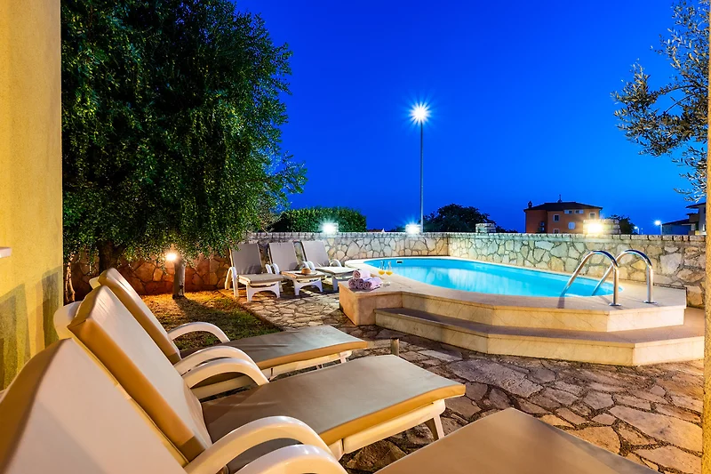 Beleuchteter Pool, Liegen und Villa im nächtlichen Ambiente.