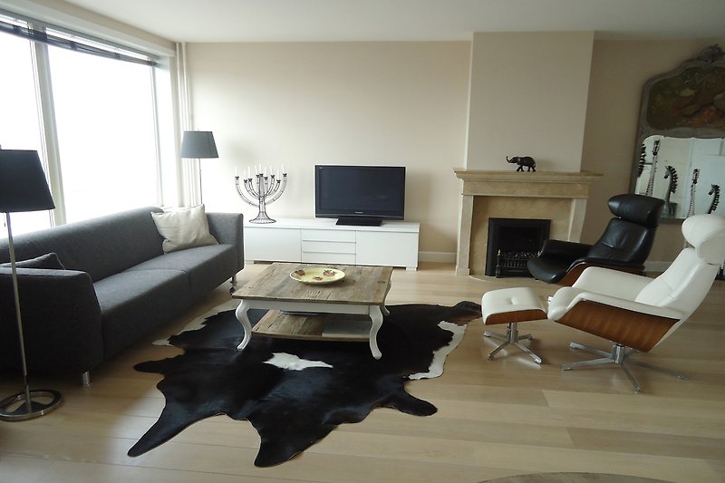 Gemütliches Wohnzimmer mit bequemer Couch, Tisch und Fernseher. Hund und Pflanzen sorgen für Atmosphäre.