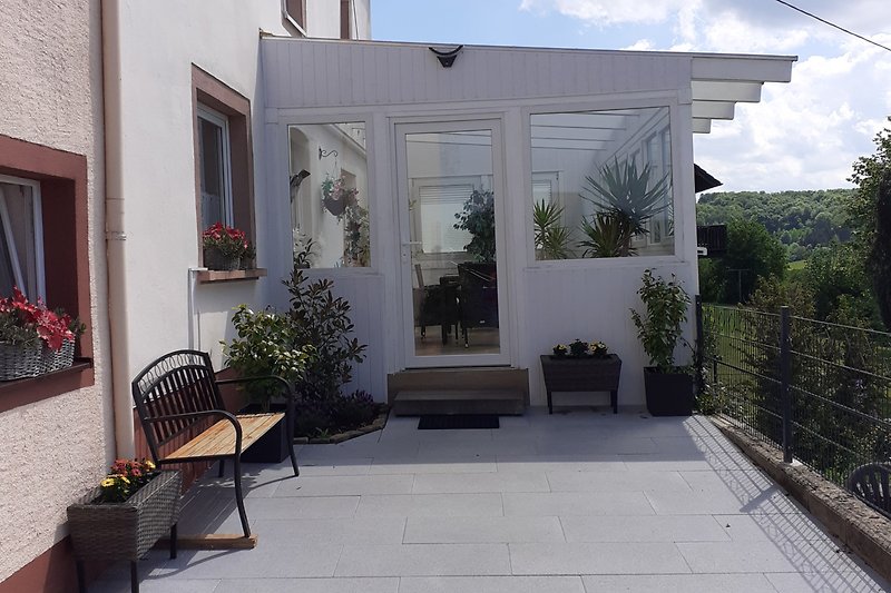 Haus mit Sauna,  Garten, Blumen, Bäumen und Terrasse unter blauem Himmel.